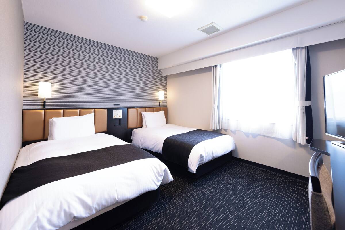 Ein Hotelzimmer mit zwei Betten, einem großen Fenster mit Vorhängen, einem Flachbildfernseher und einer Wandbedieneinheit.