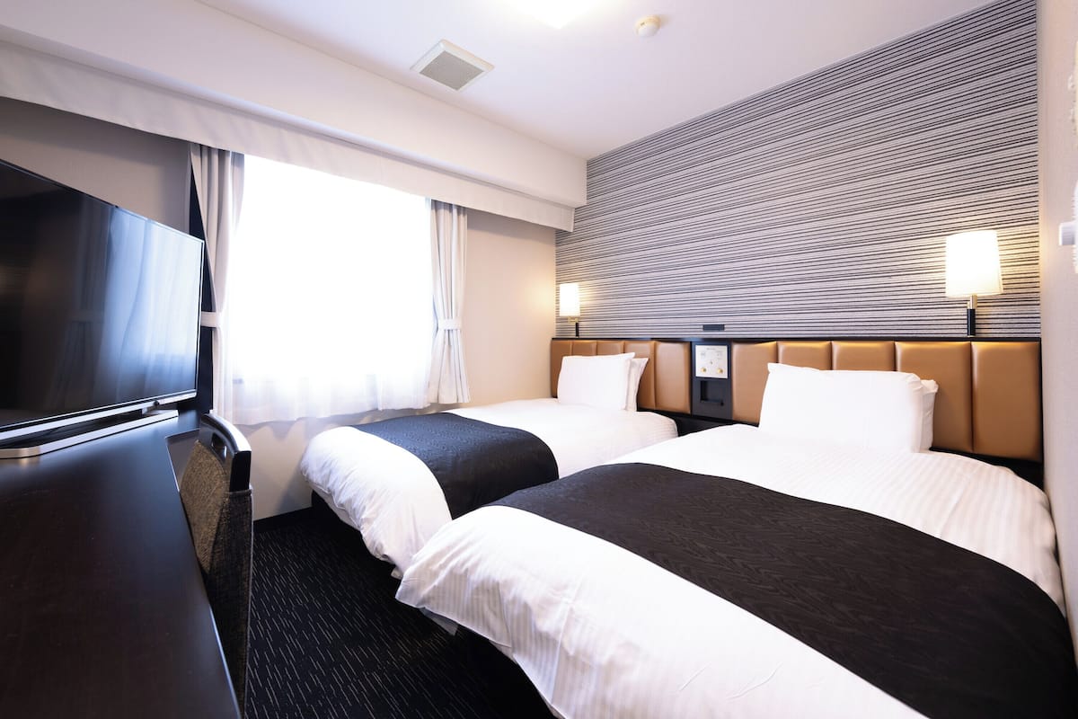 Ein Hotelzimmer mit zwei Betten, einem Fernseher, einem Schreibtisch und einem fensterbepflanzten Fenster.