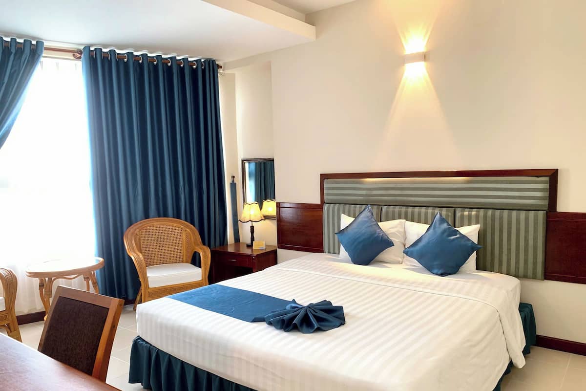 A hotel room with a large bed, a wooden nightstand, a chair, a desk, and a window with curtains.
