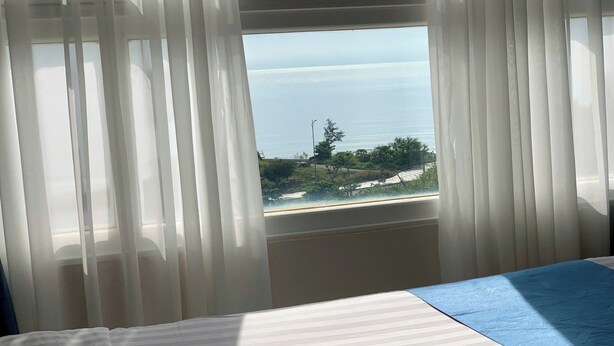 A room with a bed, a window with sheer curtains, and a view of the sea and greenery. - The Coast Hotel Vung Tau (Ho Chi Minh City)