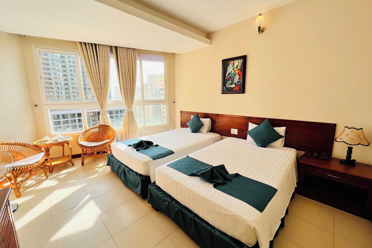 A hotel room with two beds, a sitting area with a chair and table, a painting on the wall, and a lamp on the nightstand.