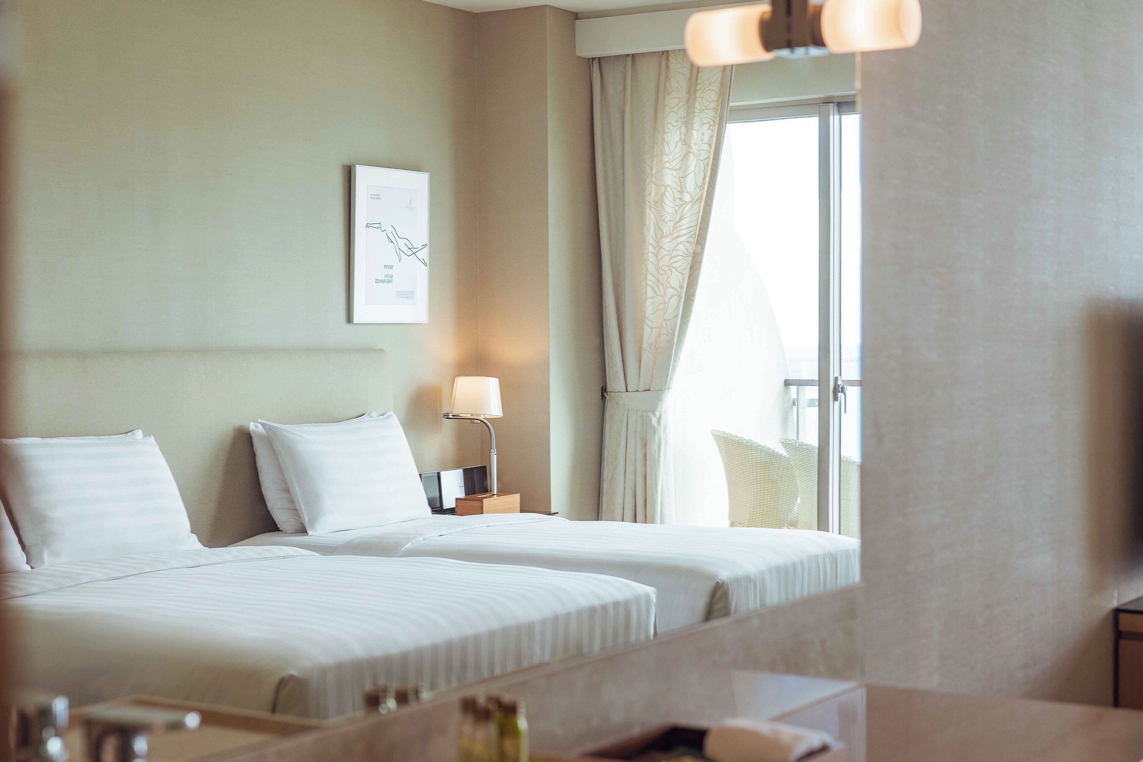 A hotel room with two beds, a bedside table with a lamp, a window with curtains, and a framed picture on the wall.