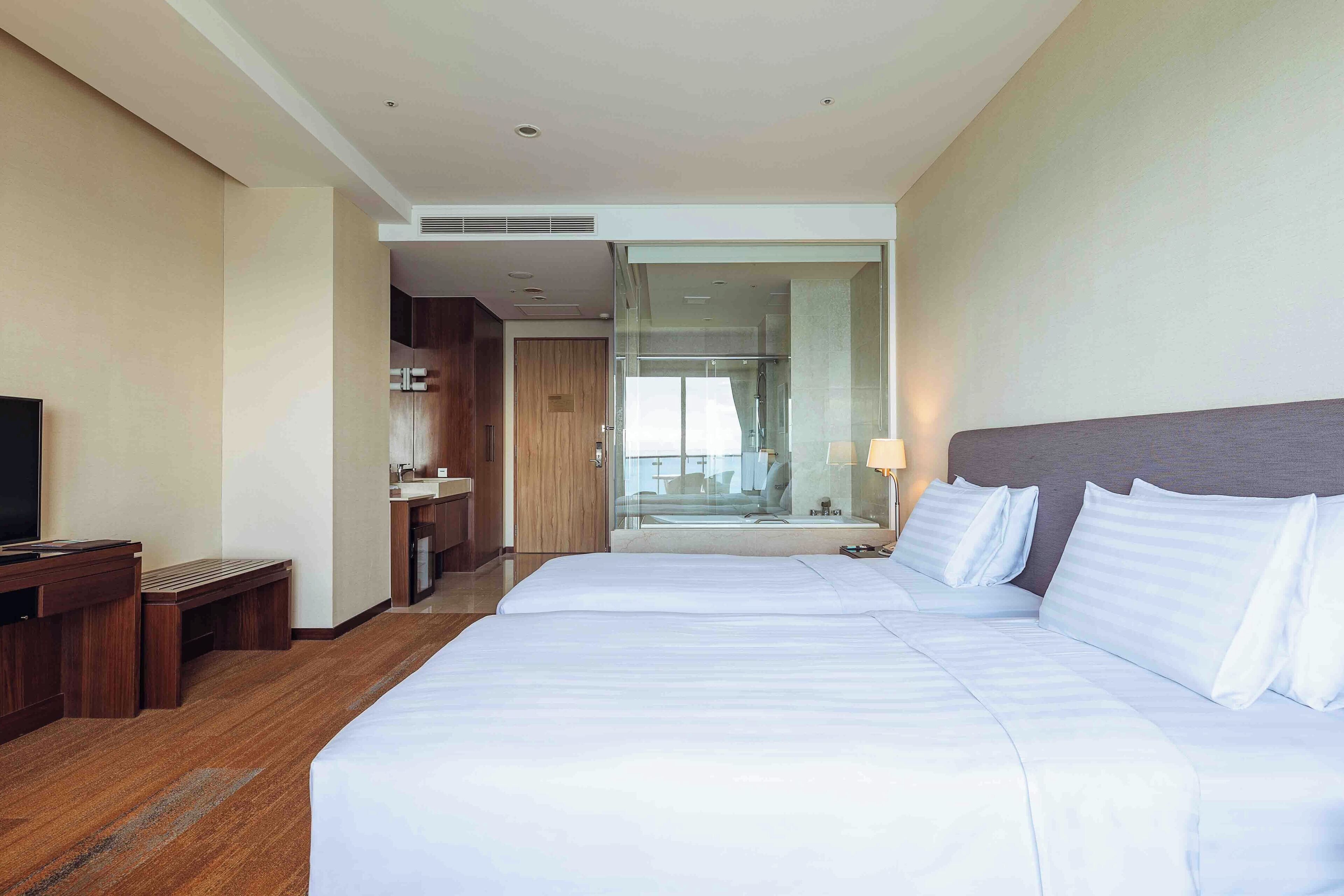 Habitación de hotel con una cama grande, televisor, escritorio y baño visible a través de una puerta de vidrio.