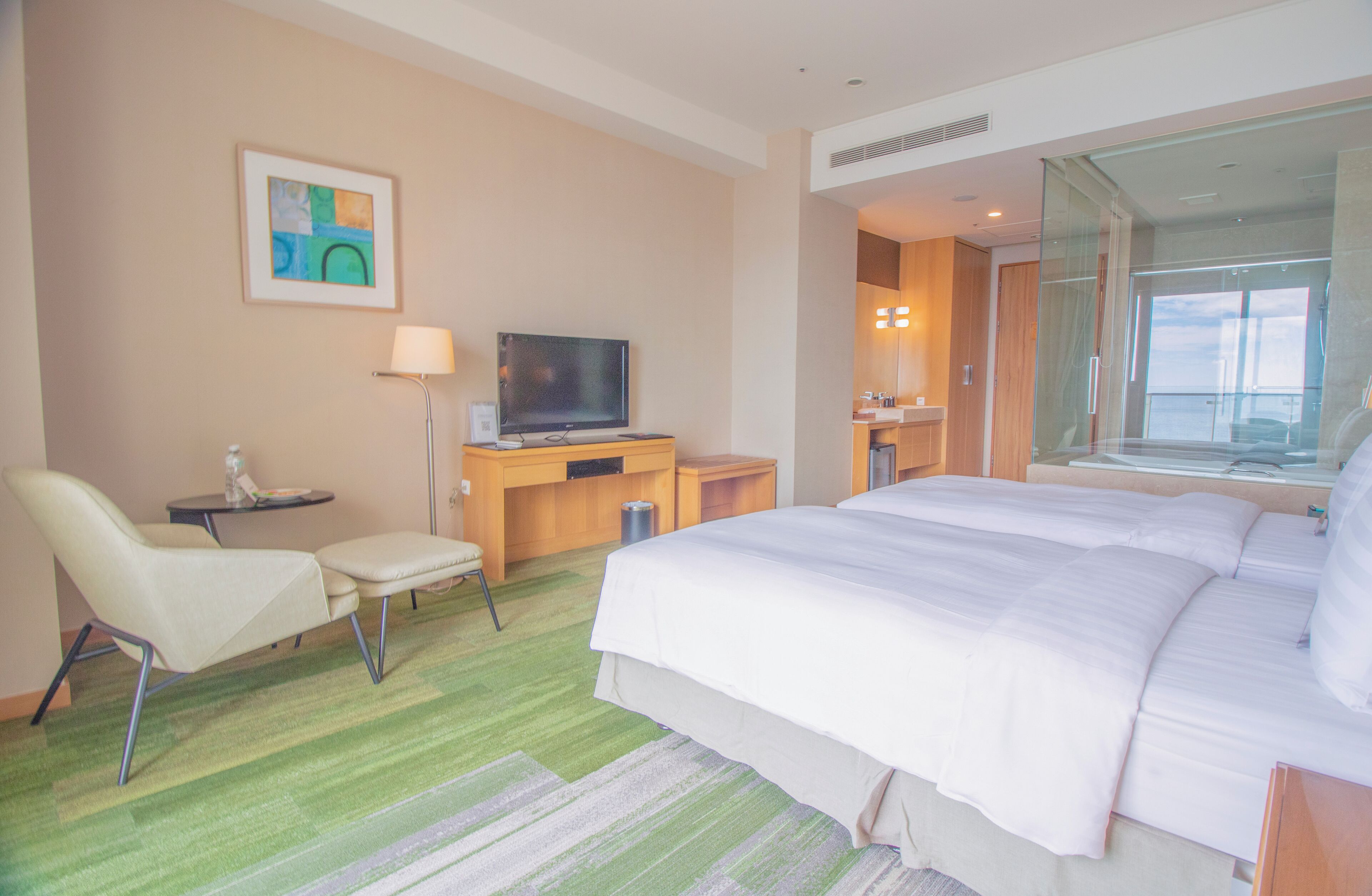 A hotel room with a large bed, a desk with a television, a chair, and a bathroom visible through a glass door.