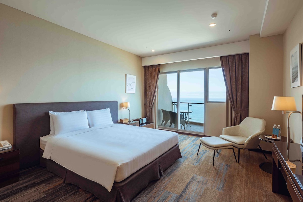 Habitación de hotel con una cama grande, un escritorio con lámpara, una silla y un balcón con vistas.