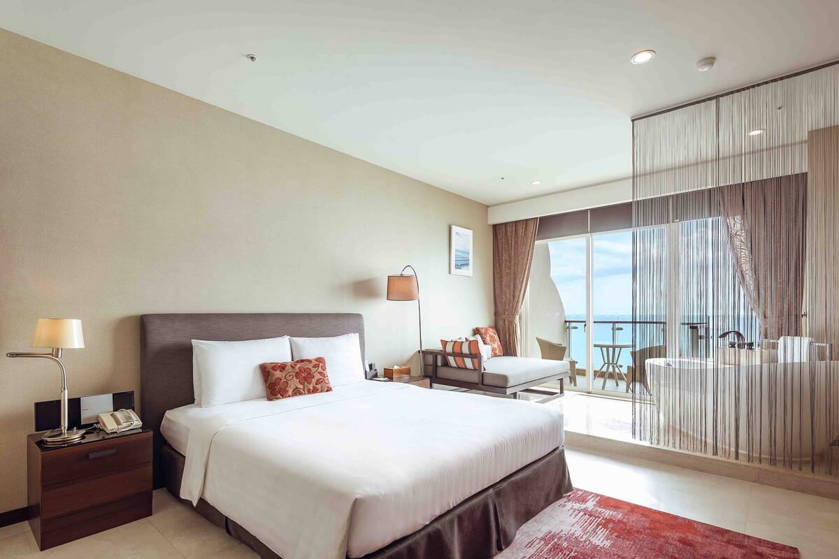 Habitación de hotel con una cama grande, un escritorio con lámpara, teléfono, una silla, una mesita y un balcón con vistas.