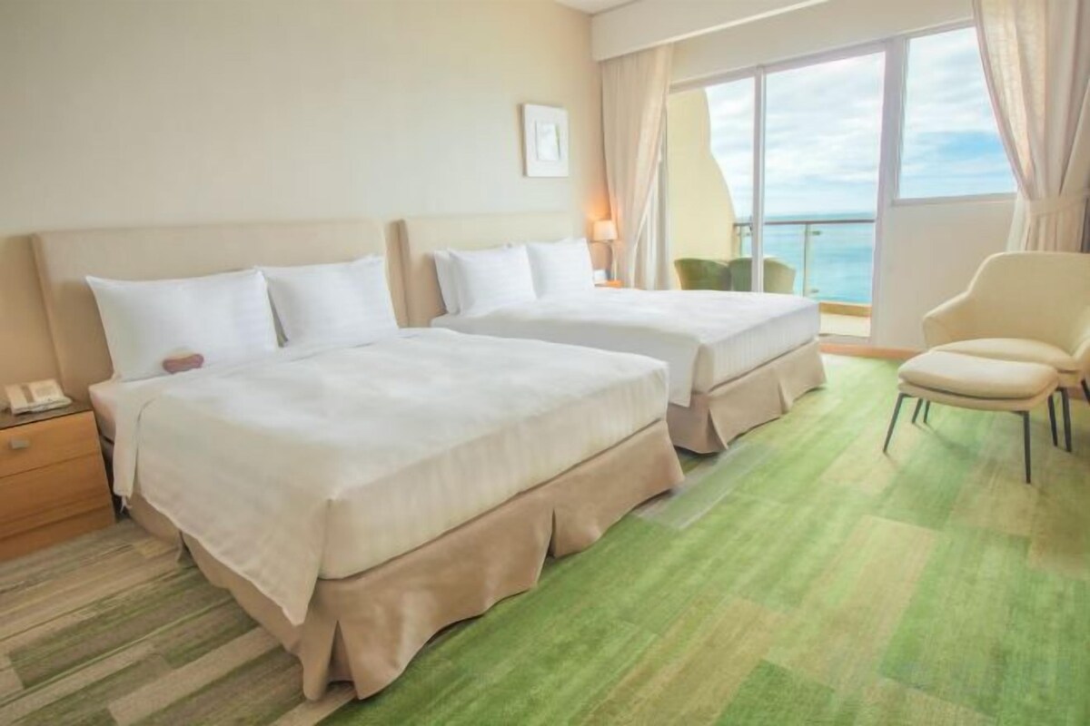 A hotel room with two beds, a chair, a nightstand, and a view of the sea.