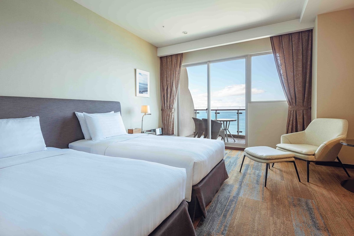 Habitación de hotel con dos camas, un balcón con mesa y sillas, y vista al mar.