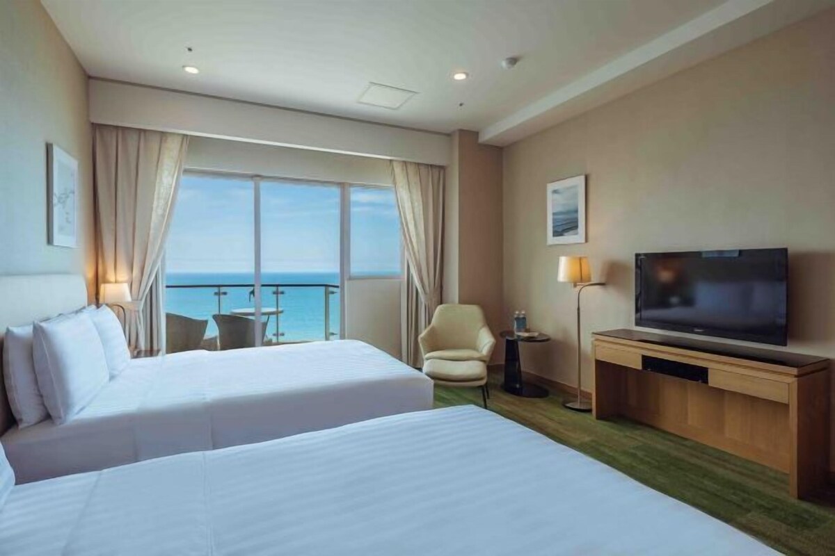 Une chambre d’hôtel avec deux lits, une télévision, une chaise et une vue sur la mer.