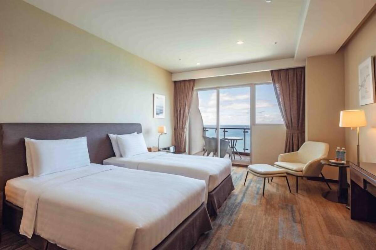 Habitación de hotel con dos camas, un balcón con mesa y sillas, y un escritorio con lámpara.