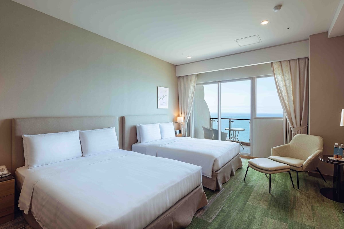 Habitación de hotel con dos camas, una silla, una mesita y vista al mar.