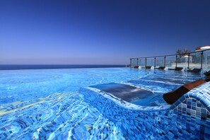 Una piscina infinita con vista despejada al océano, barandilla de cristal y sillas de descanso.