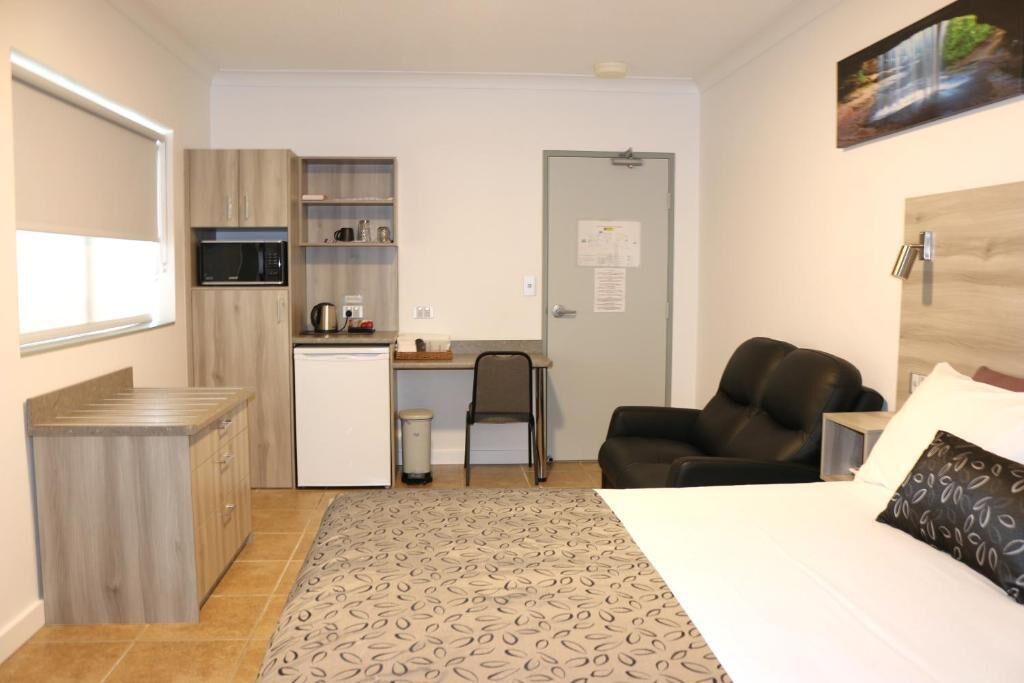 A compact hotel room with a bed, a desk with a chair, a kitchenette, and a door.
