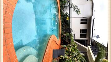 A swimming pool with a brick border, surrounded by potted plants and a building with a satellite dish.