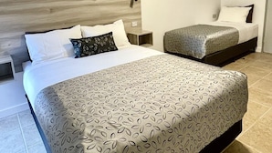 Une chambre d’hôtel avec un grand lit, un lit plus petit, une tête de lit en bois, une table de chevet et un tableau accroché au mur.