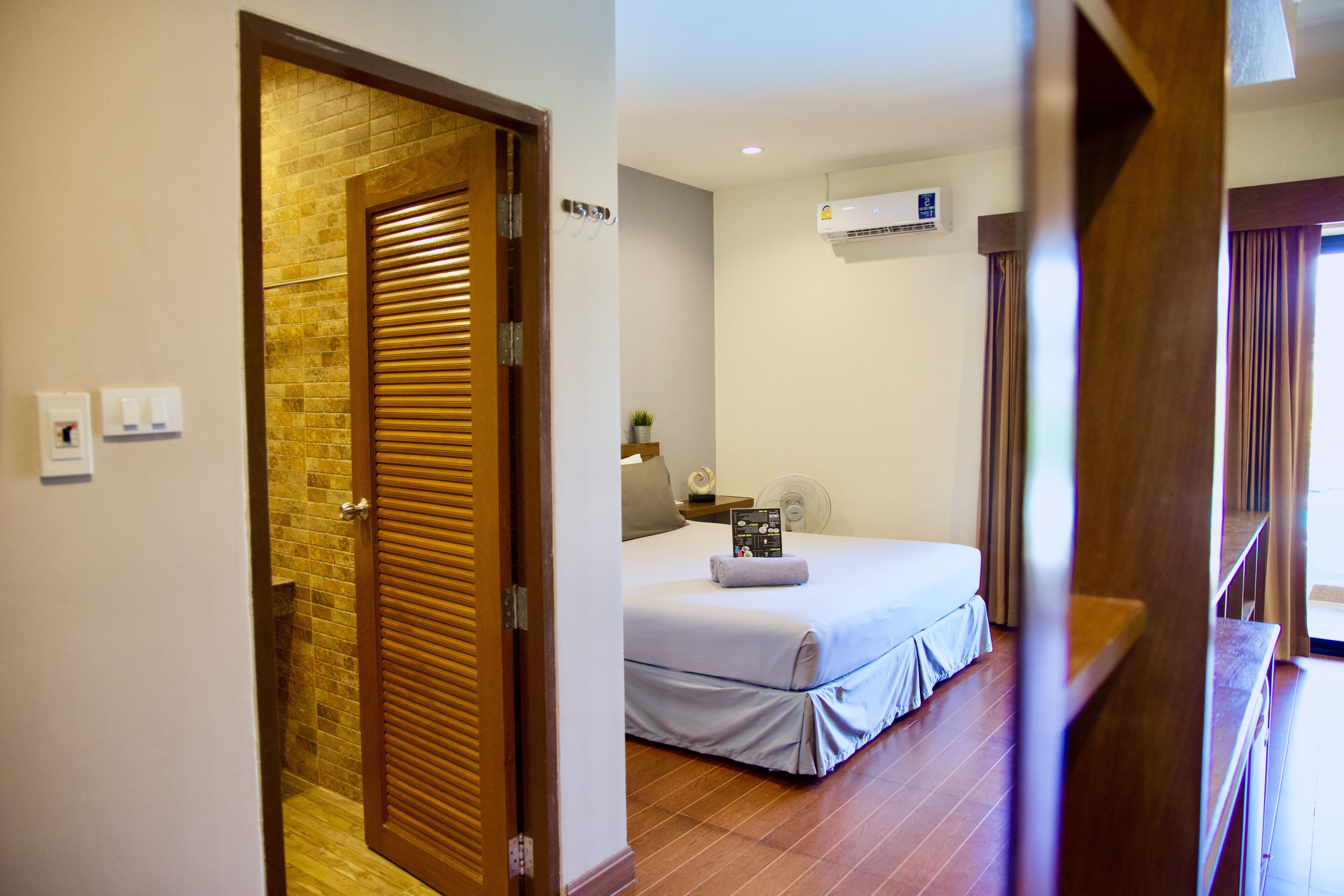 A hotel room with a wooden door, a bed with a white bedspread, a bedside table with a lamp, and an air conditioning unit on the wall.