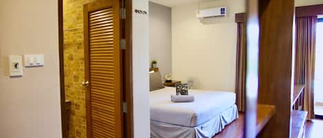 A hotel room with a wooden door, a bed with a white bedspread, a bedside table with a lamp, and an air conditioning unit on the wall.