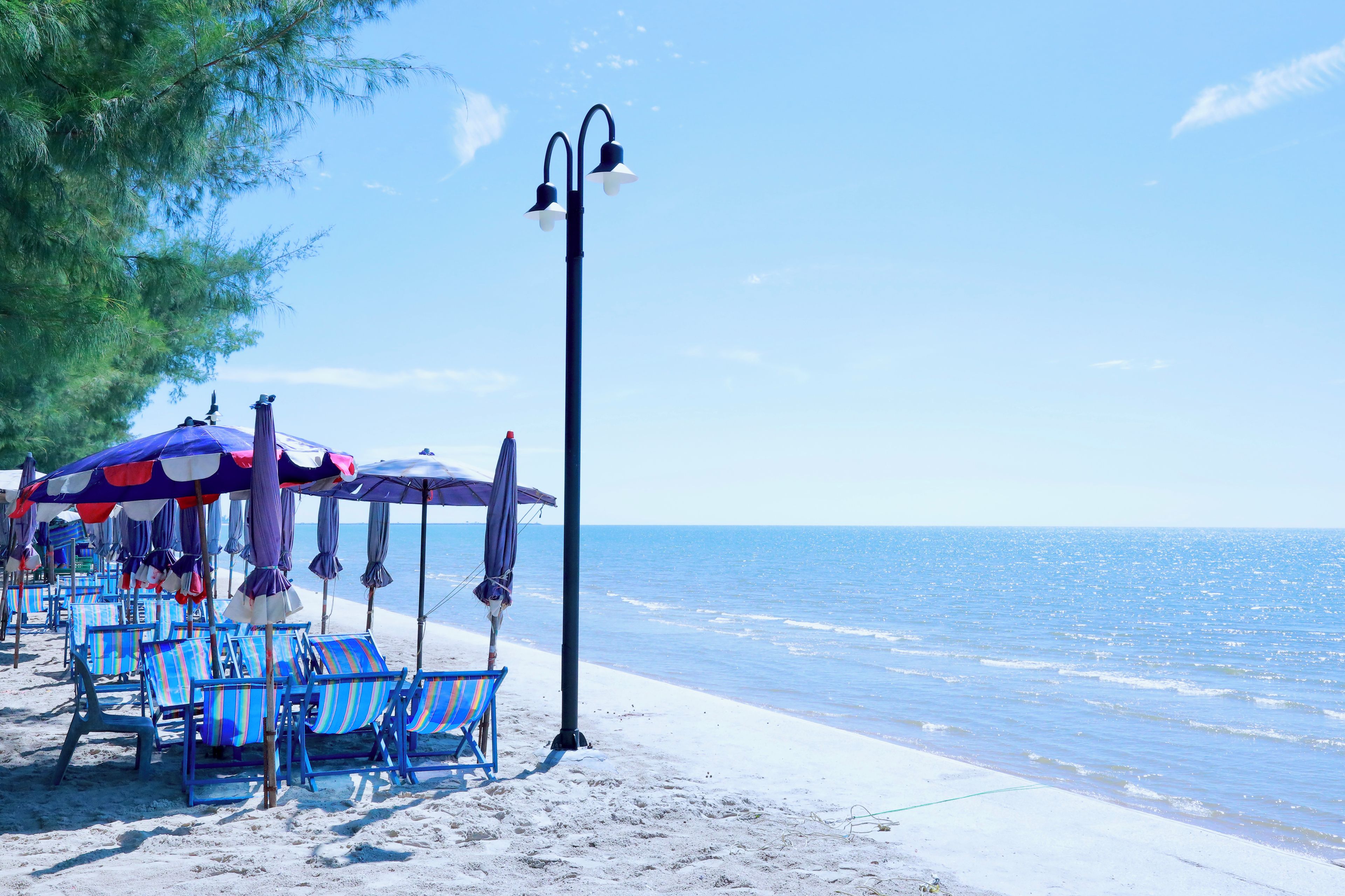 A beach with rows of striped beach chairs and umbrellas, a tall street lamp, and a clear view of the sea.