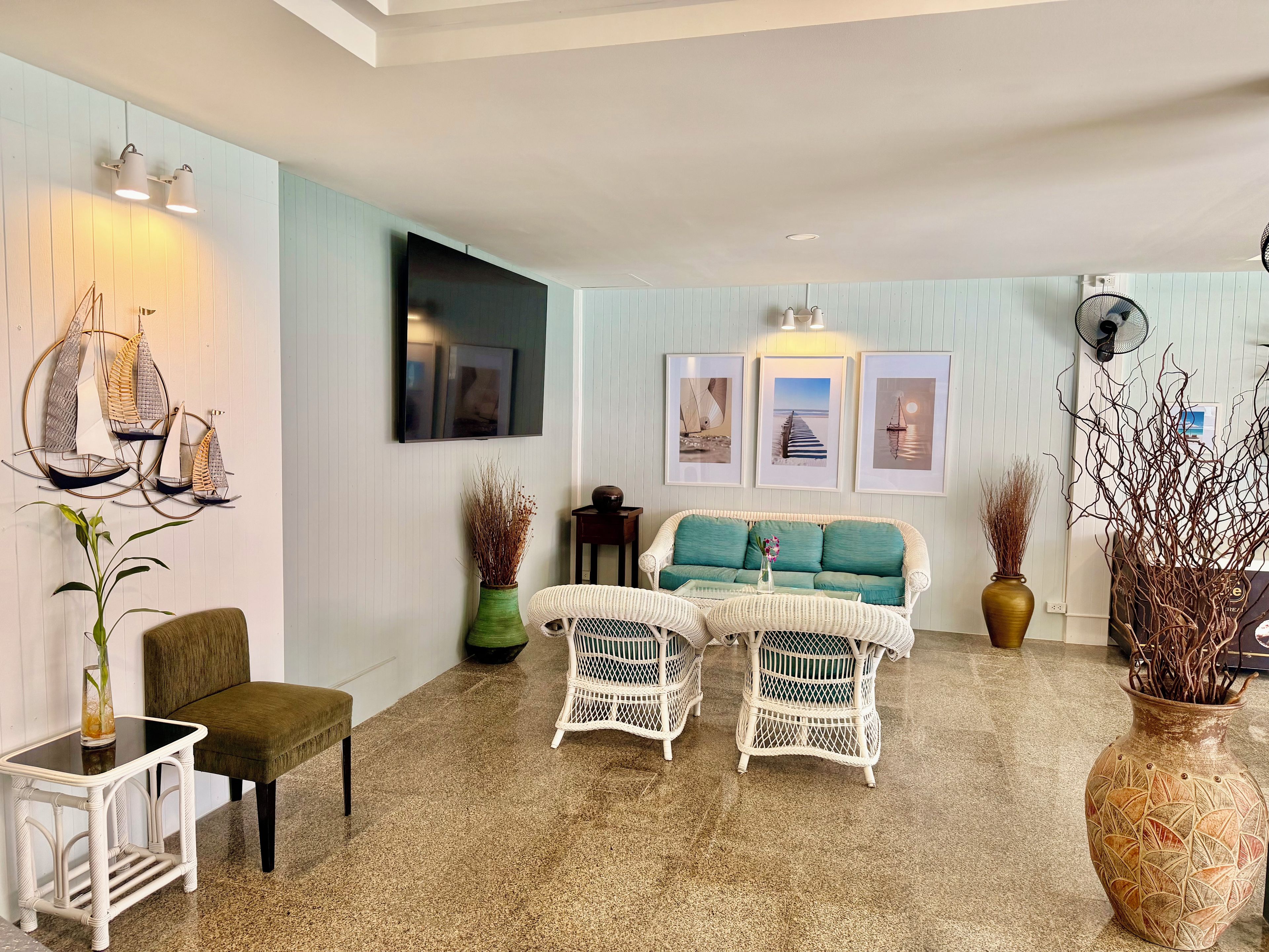 A living room with a sofa, two white wicker chairs, a small table, a TV, wall-mounted artwork, and a fan.