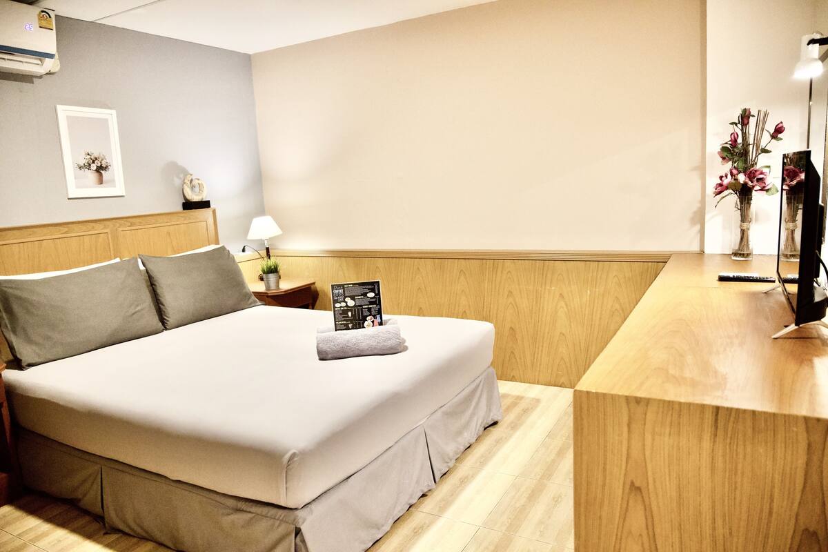 A neatly made bed with a grey bedspread and pillows, a wooden headboard, a bedside table with a lamp, a framed picture, and a vase with flowers on the right side.