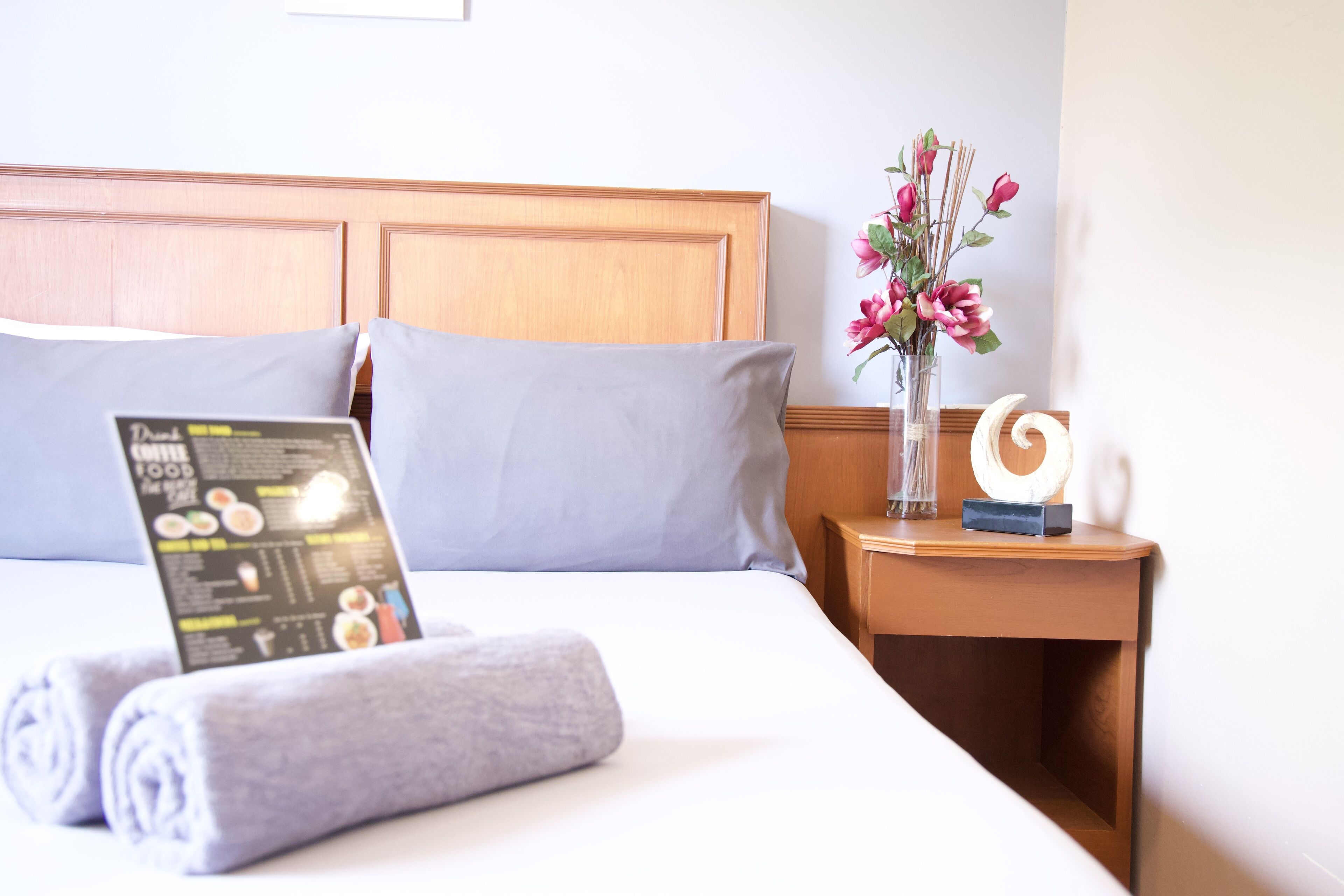 A neatly made bed with a pillow, a rolled towel, a menu, a vase with flowers, and a decorative item on a wooden nightstand.