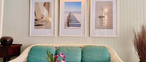 A white wicker sofa with teal cushions, a vase with flowers, and three framed pictures on the wall.