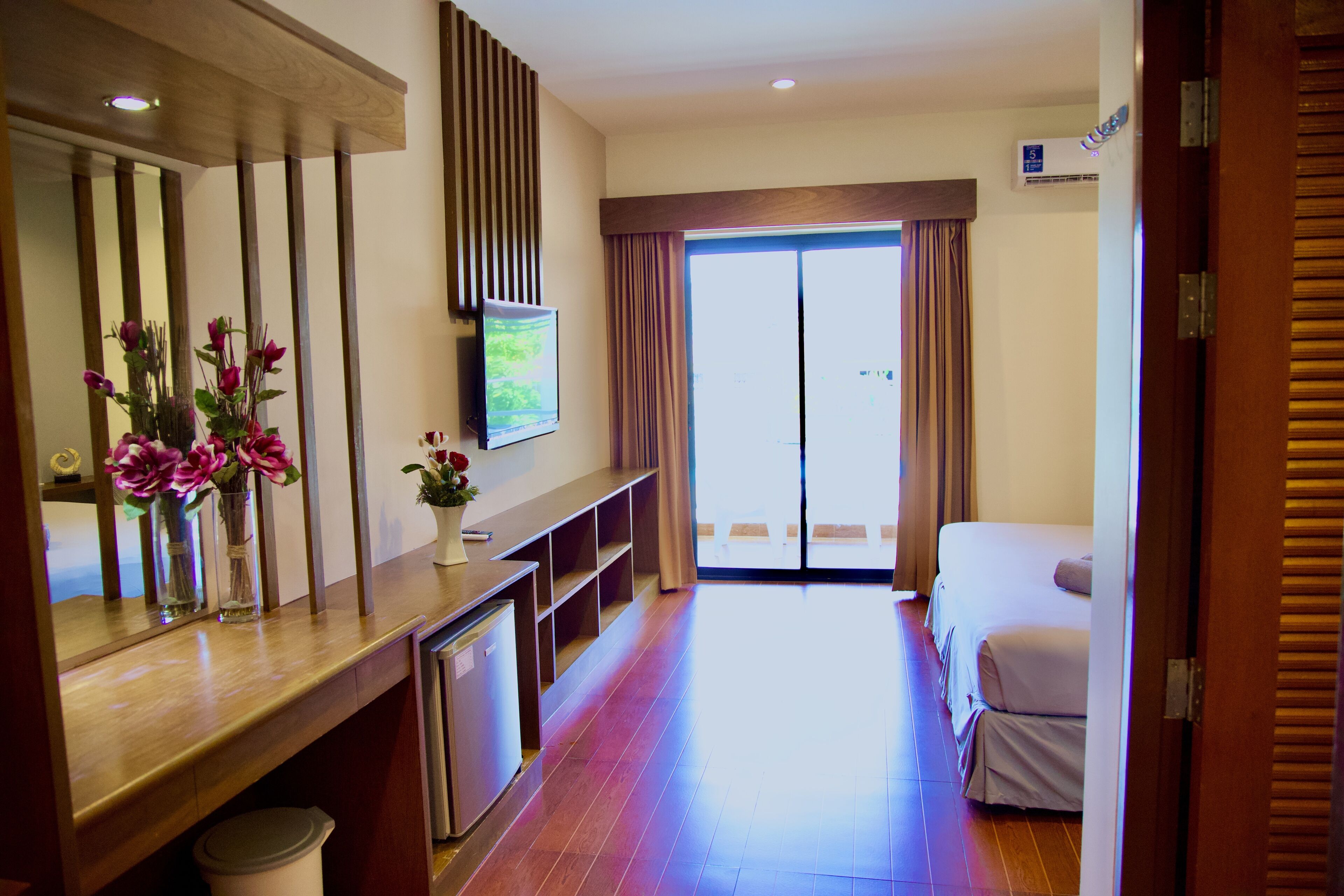 A hotel room with a wooden desk, a television, a bed, and a sliding glass door leading to a balcony.