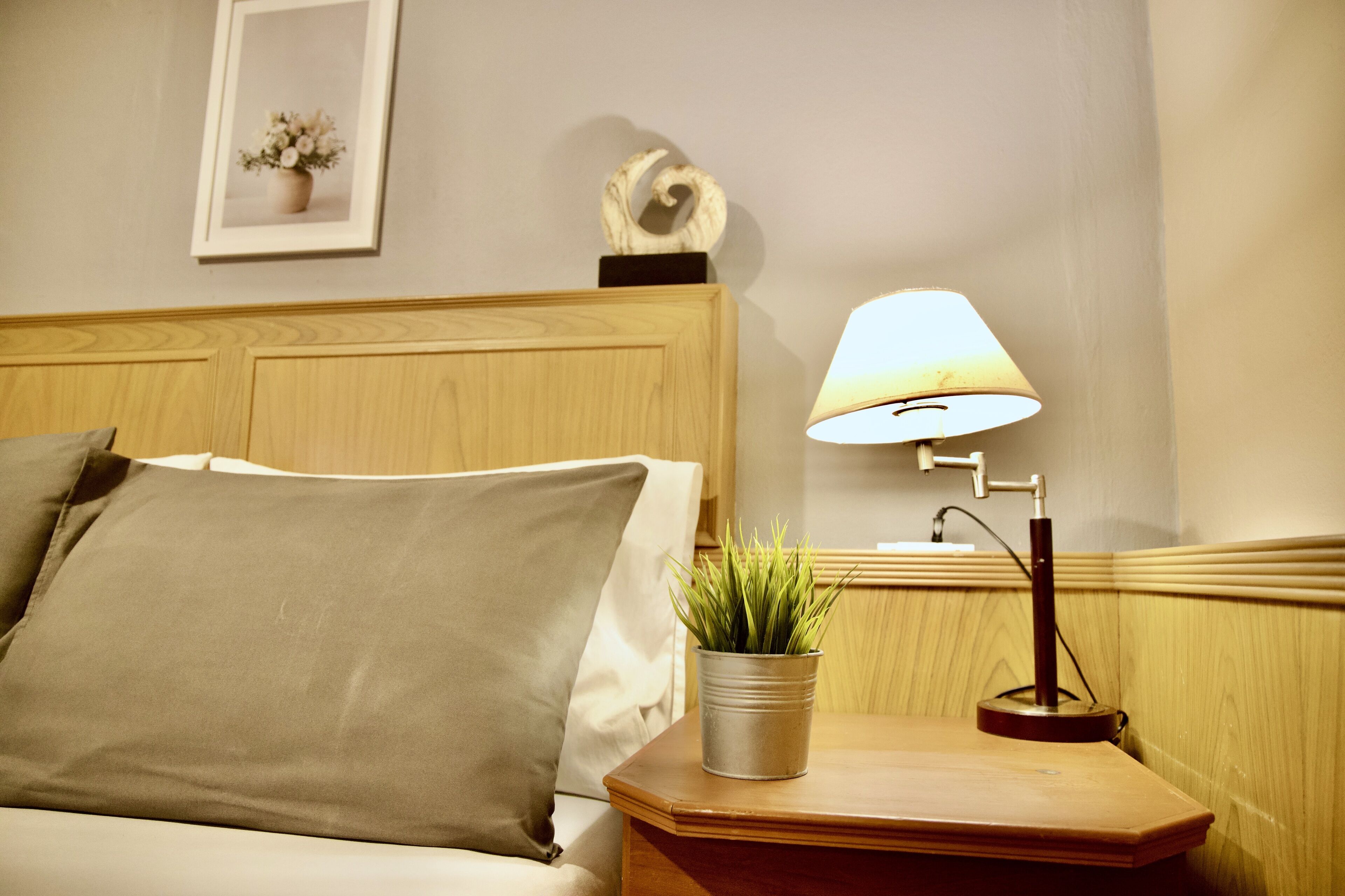 A neatly made bed with a pillow, a bedside table with a lamp, a small potted plant, and a framed picture on the wall.