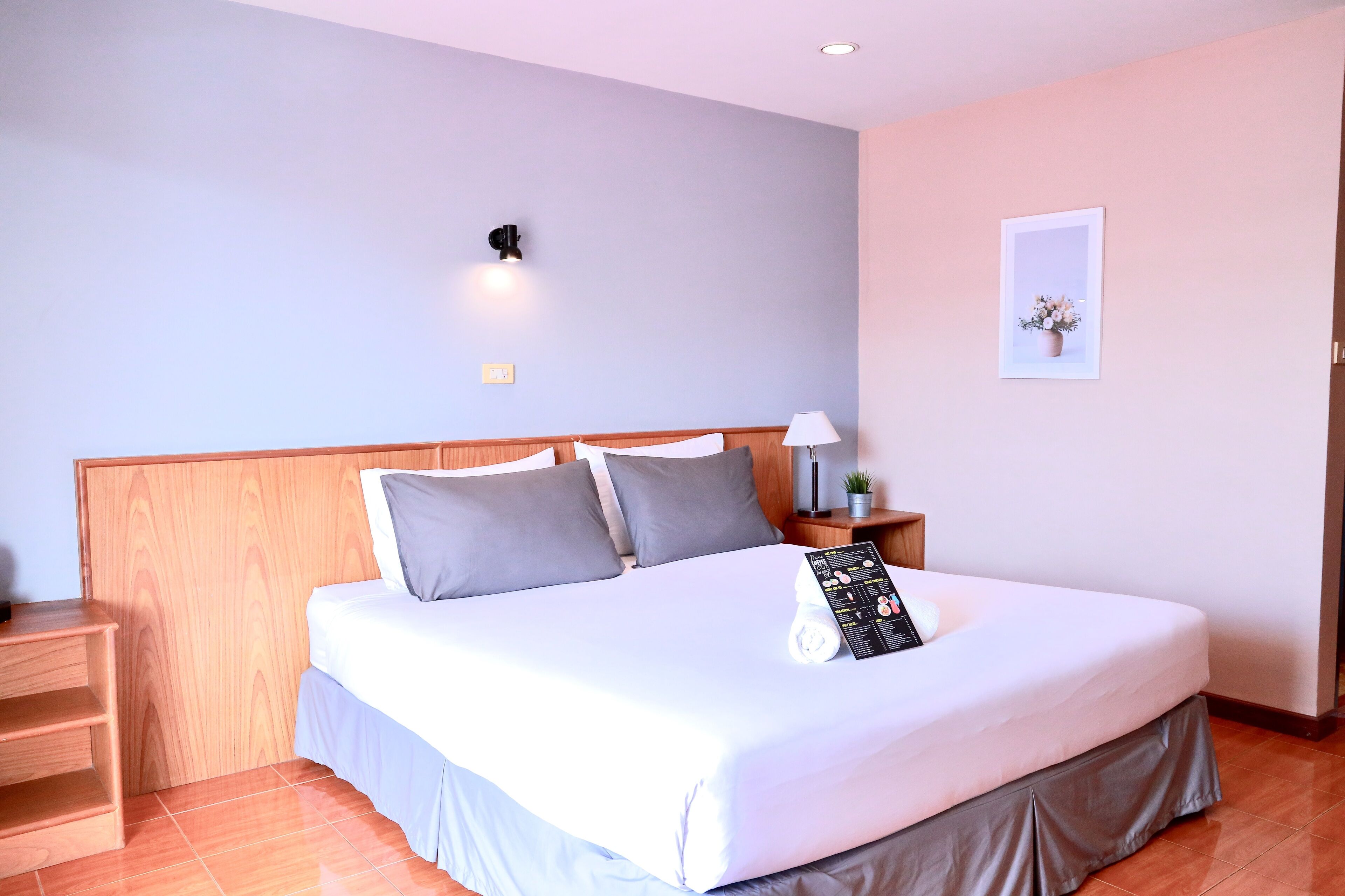 A neatly made bed with a white comforter and gray pillows, a wooden headboard, a nightstand with a lamp, and a framed picture on the wall.