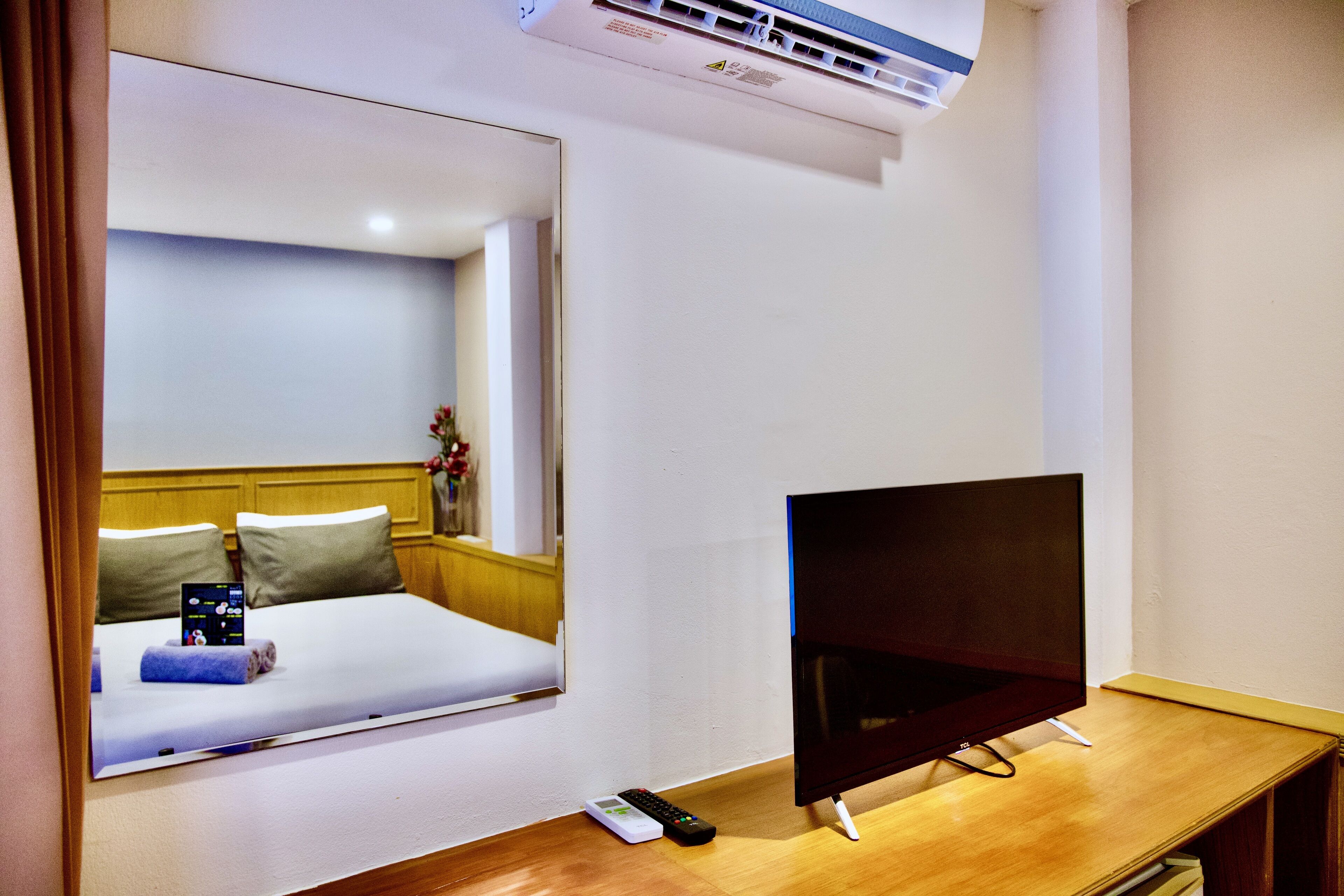 A hotel room with a bed, a TV on a wooden stand, a remote control, and an air conditioning unit.