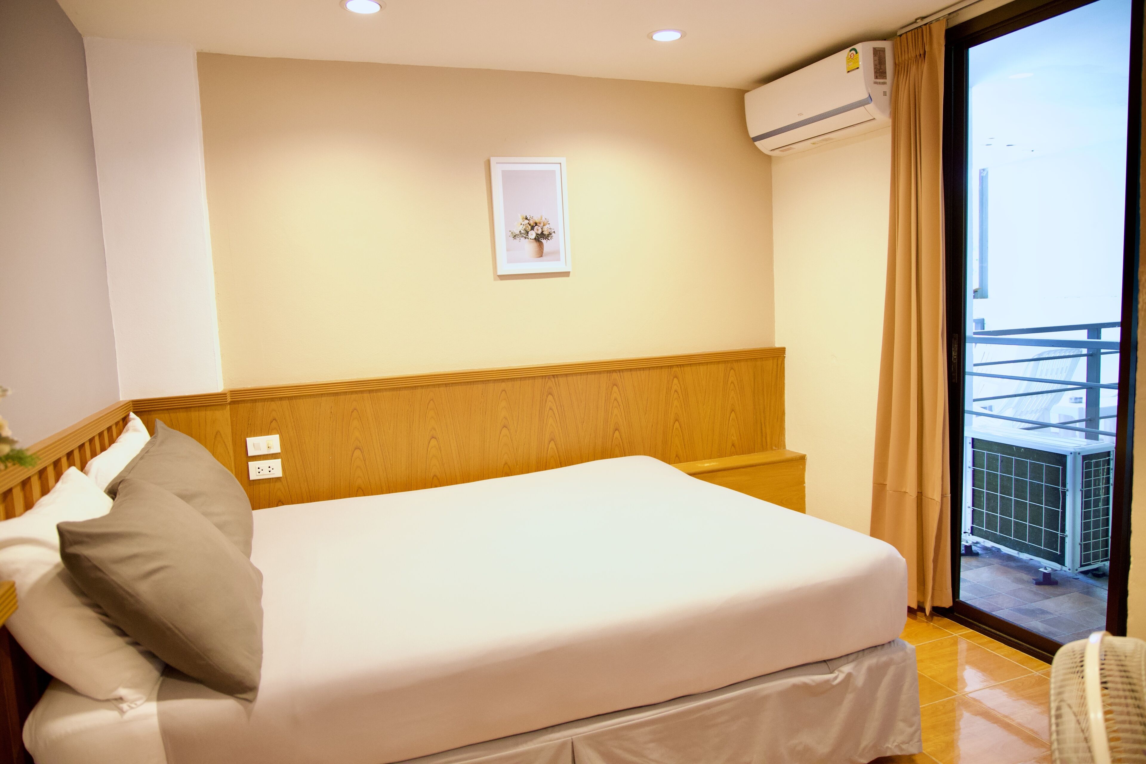 A hotel room with a bed, a small bedside table, a wall-mounted air conditioner, a framed picture, and a view of the outside balcony.