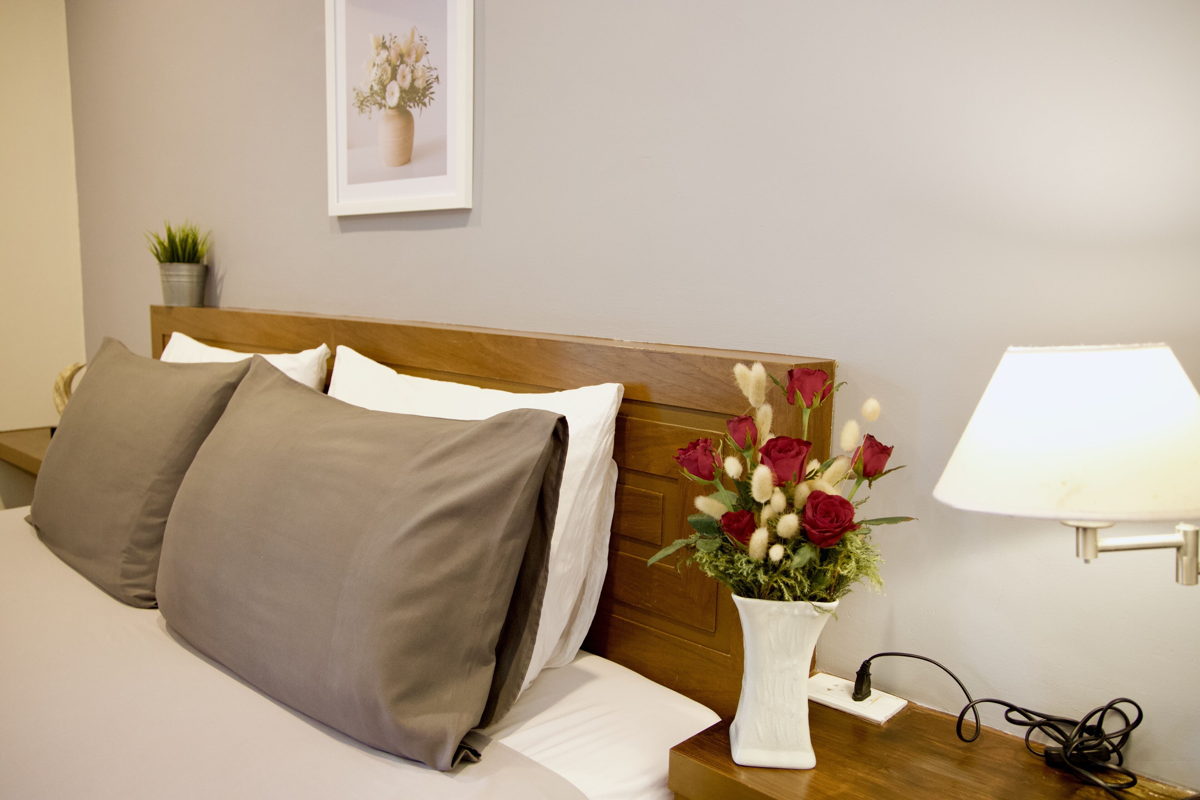 A neatly made bed with grey and white linens, a wooden headboard, a white vase with red roses, and a bedside lamp on a wooden nightstand.
