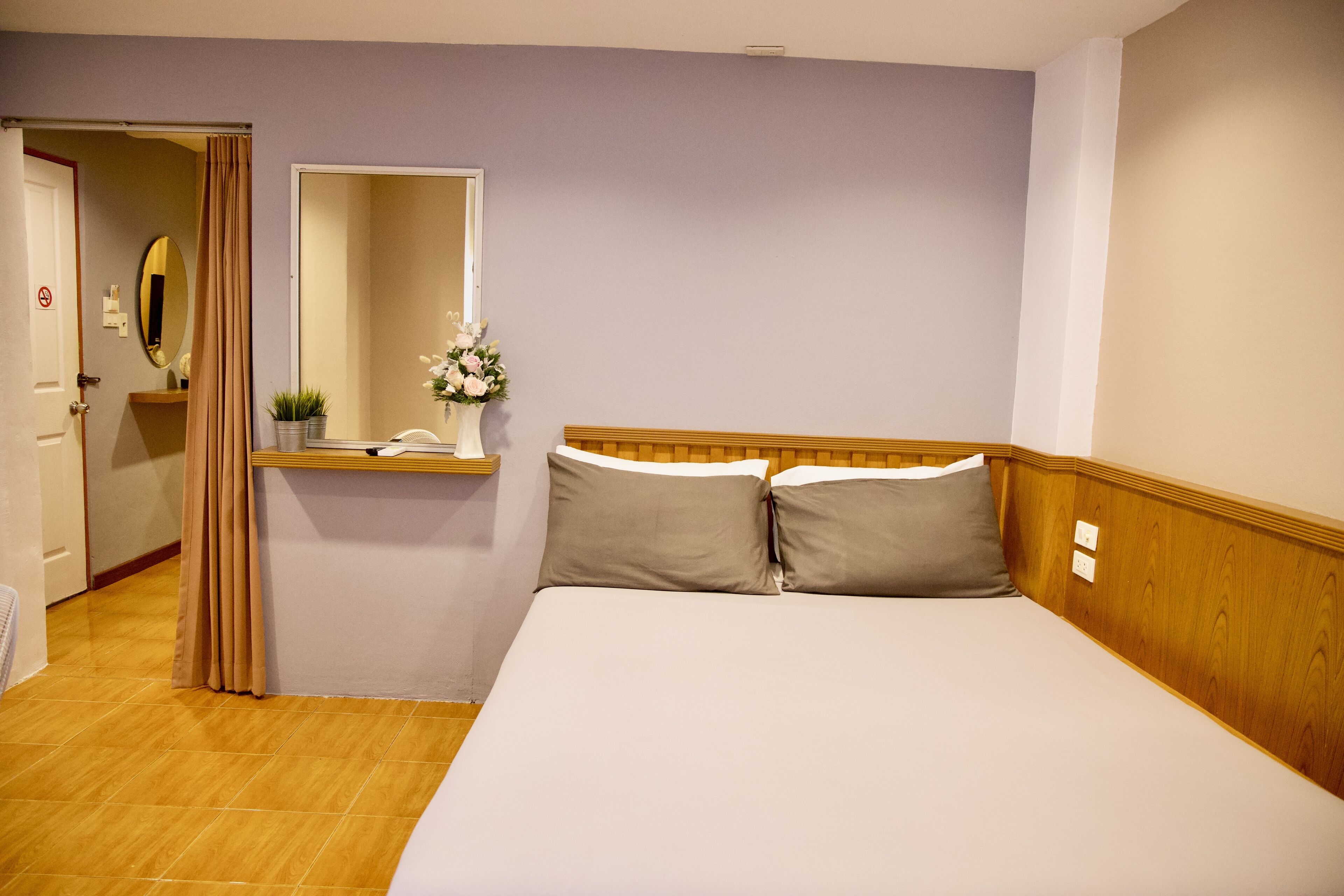 Une chambre d’hôtel avec une tête de lit en bois, un lit avec deux oreillers, une table de chevet avec un miroir, une petite plante en pot et une porte donnant sur une autre pièce.