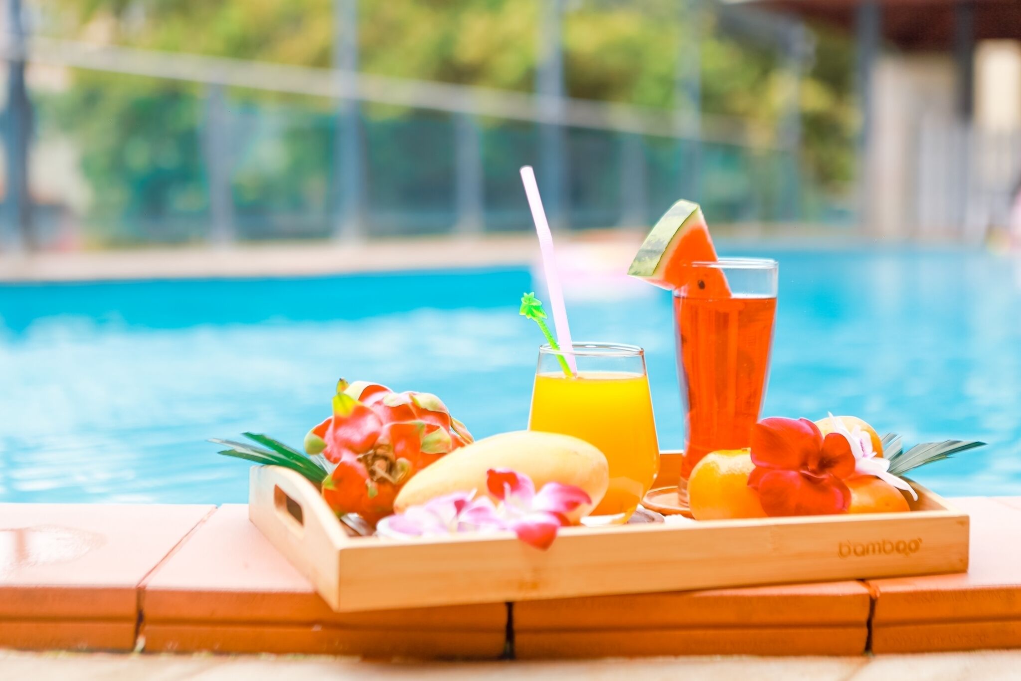 A wooden tray with a glass of orange juice, a tall glass of orange beverage, and a banana, placed by a poolside.