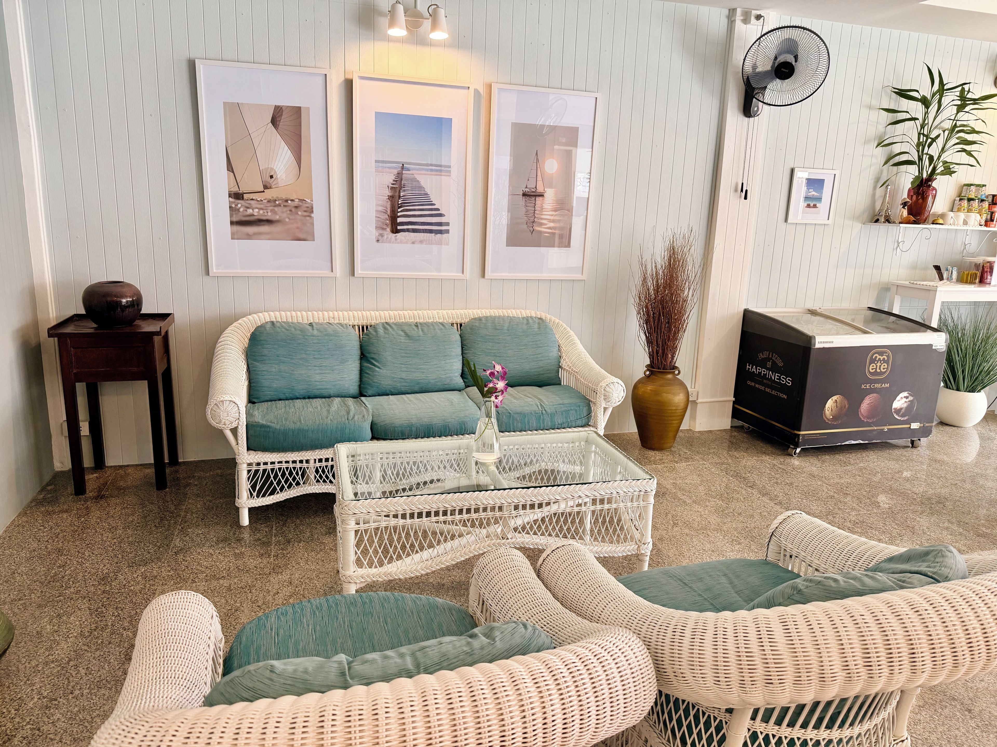 A living room with a white wicker sofa, a coffee table, and a fan. There are framed pictures on the wall, a vase with dried plants, and a potted plant.