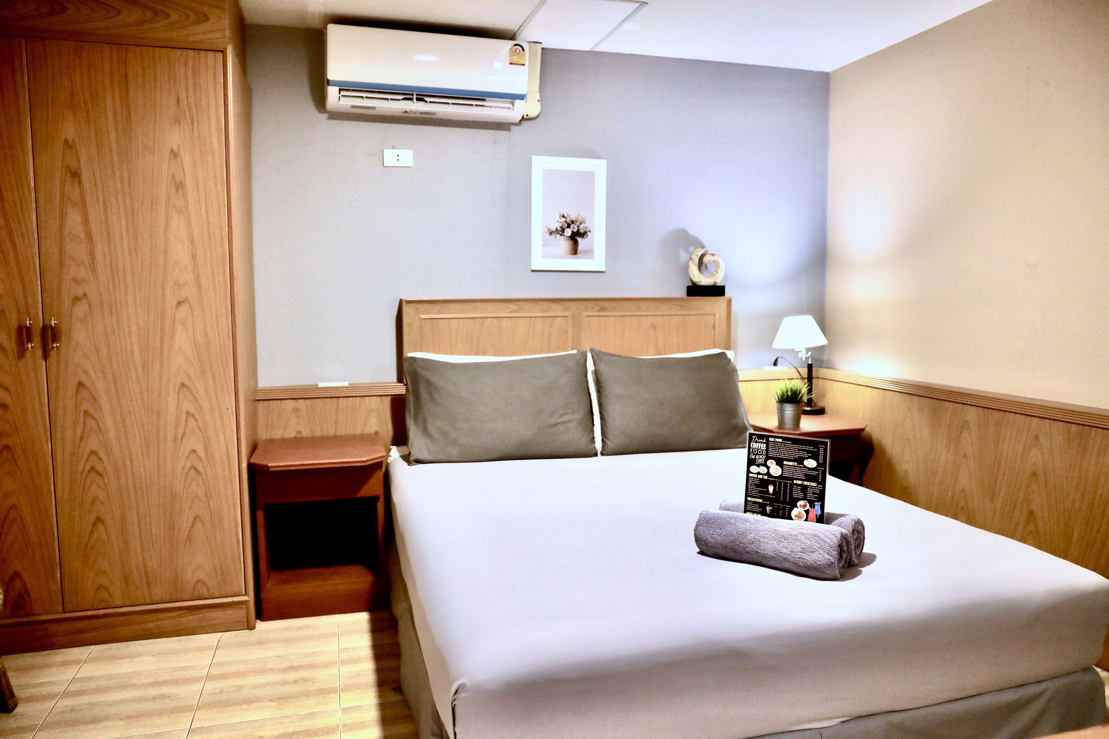 A hotel room with a wooden wardrobe, a bed with a headboard, a nightstand, a lamp, and a framed picture on the wall.