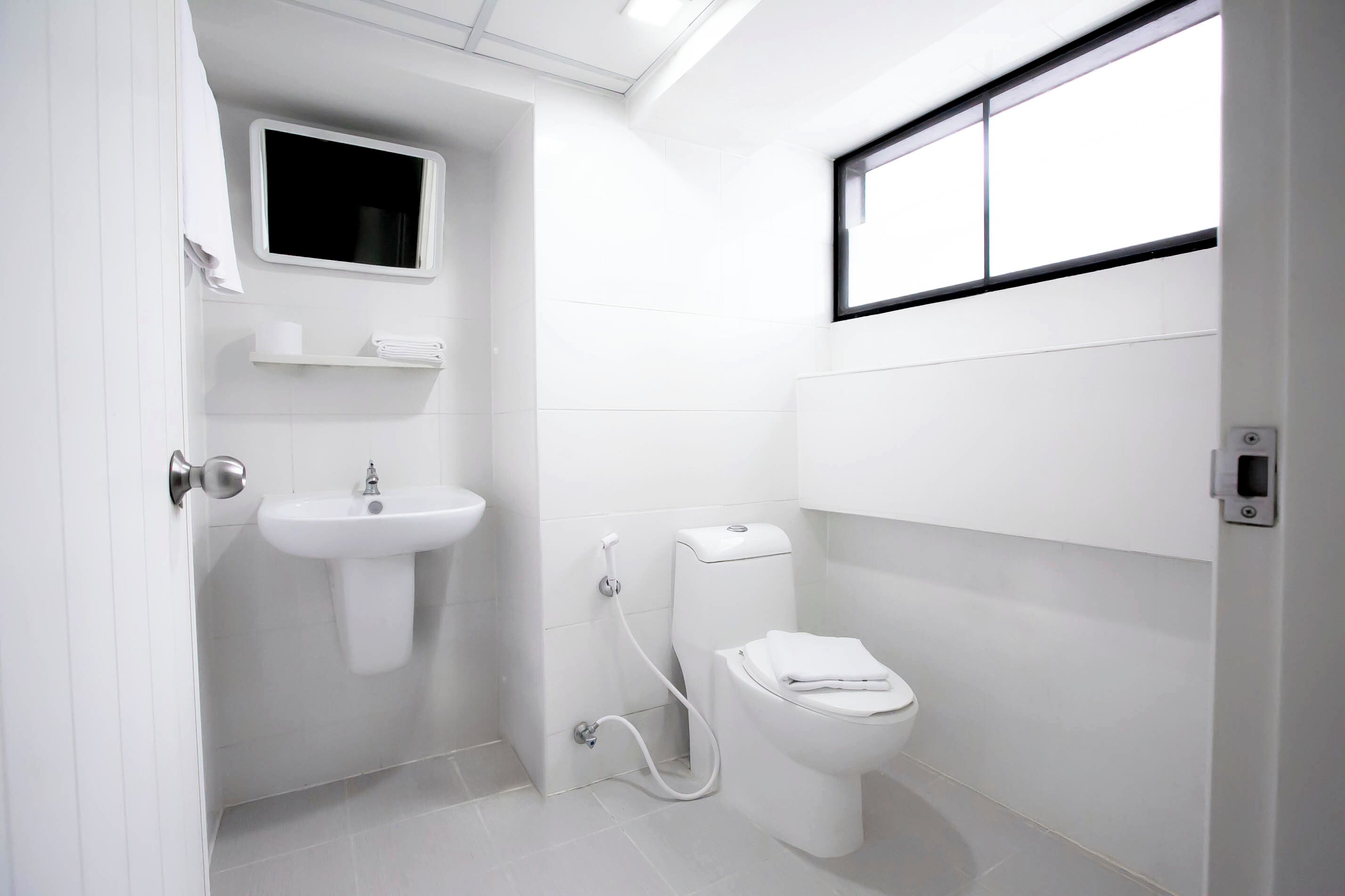 A modern bathroom with a white sink, toilet, and shower. There is a window above the toilet and a small shelf with towels.
