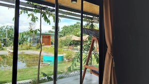 A modern room with large glass windows, a view of a garden with a pond, and a swing set. - Ban Bang Home Resort (Phetchaburi)