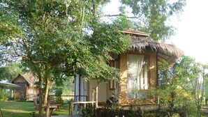 A rustic cabin with a thatched roof, surrounded by lush greenery and a small garden. - Ban Bang Home Resort (Phetchaburi)