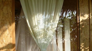 A room with a wooden wall, a window with a sheer curtain, and a view of trees outside. - Ban Bang Home Resort (Phetchaburi)