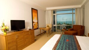 A hotel room with a bed, a TV, a wooden cabinet, a small table with a vase of flowers, a chair, and a large window with a view of the sea. - Petro Hotel (Ho Chi Minh City)