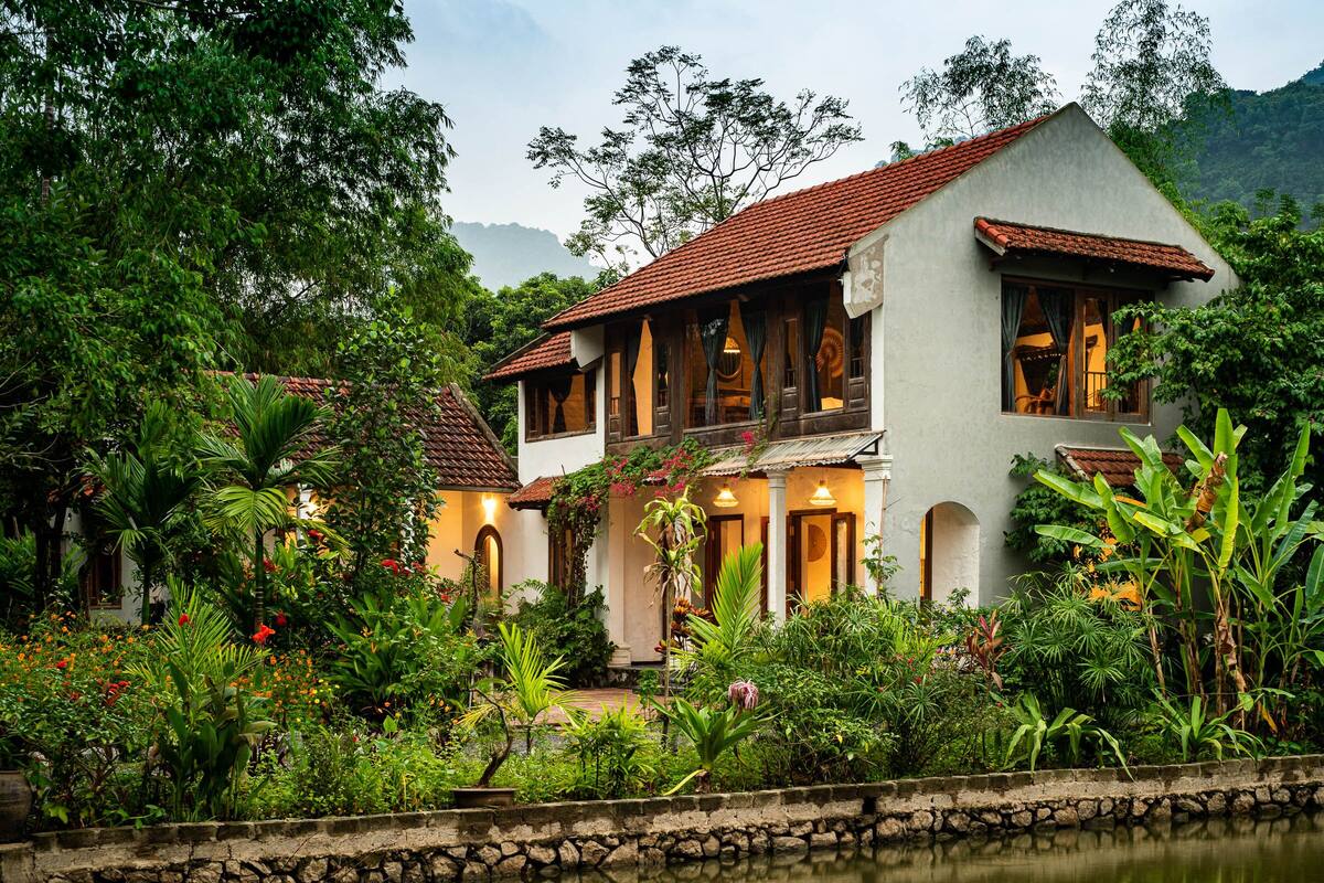 A two-story house with a tiled roof, surrounded by lush greenery and a water feature in the foreground.