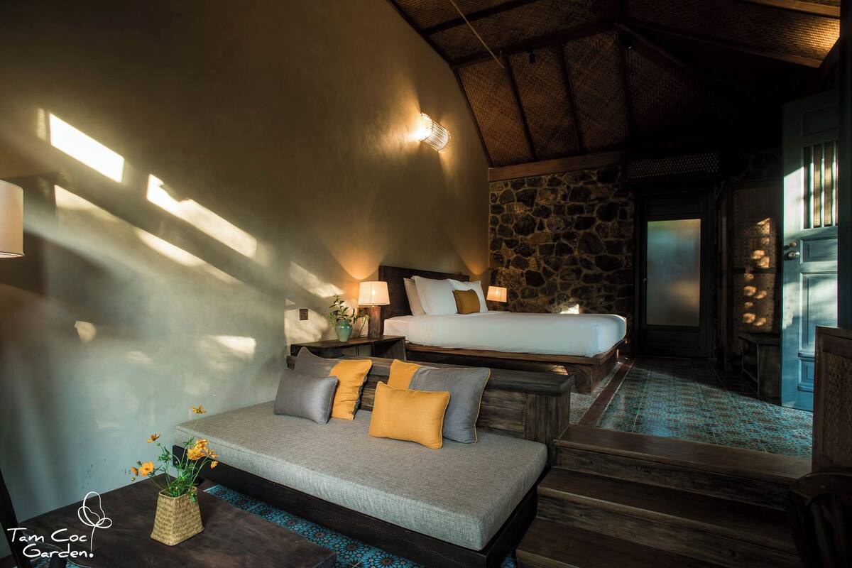 A cozy room with a sofa, a bed, and a stone wall. There is a small table with a vase and a lamp.