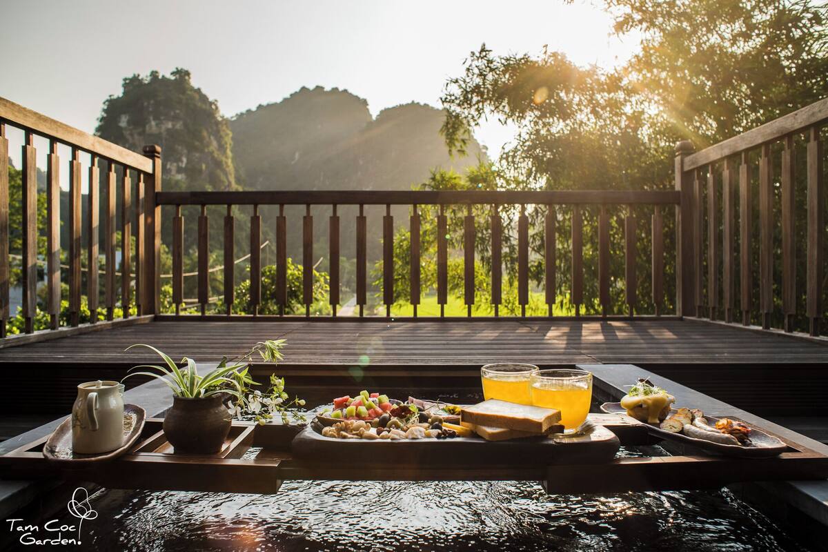 A wooden deck with a tray of food and drinks, overlooking a lush garden and mountains.