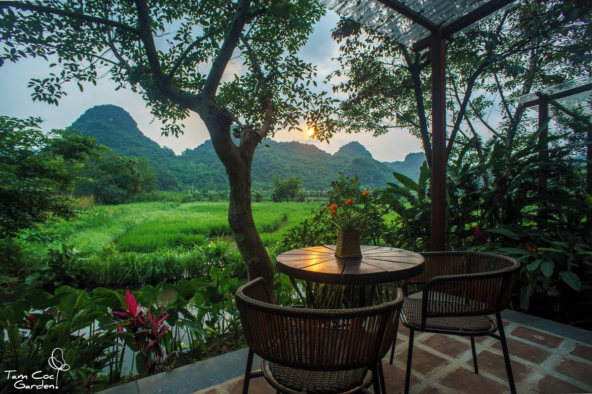 A round table with wicker chairs, a vase with flowers, and a view of a lush green landscape with hills in the background.