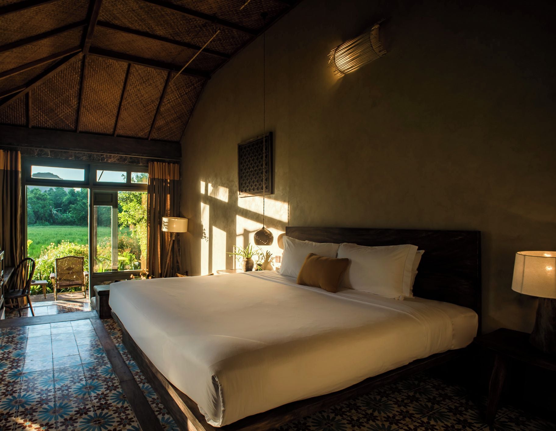 A large bed with a wooden headboard, a nightstand, a lamp, and a view of greenery outside.