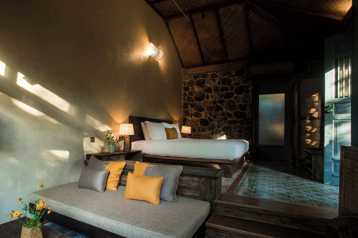 A cozy room with a sofa, a bed, and a stone wall. There is a small table with a vase and a lamp.