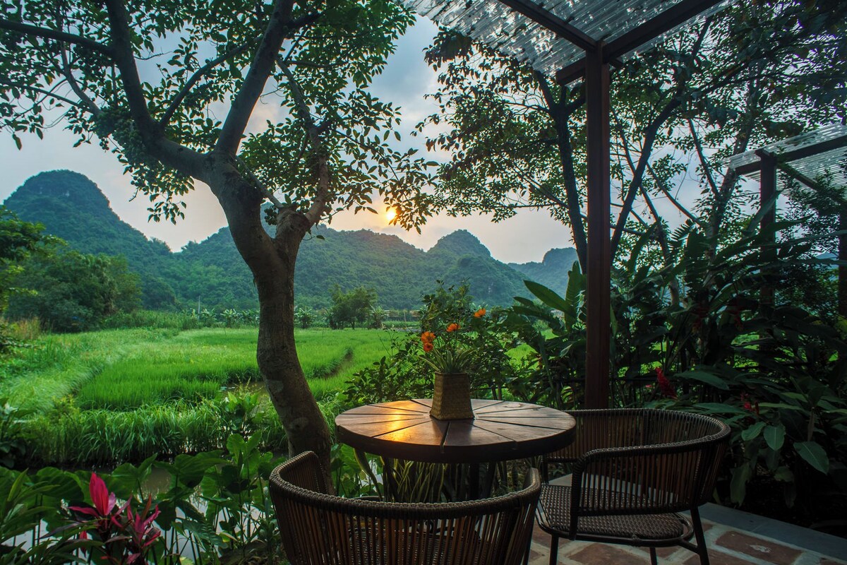 A round table with wicker chairs, a vase with flowers, and a view of a lush green landscape with hills in the background.