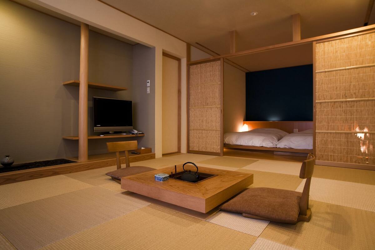 A modern hotel room with tatami flooring, a flat-screen TV, a bed with a nightstand, and a traditional Japanese-style table with a kettle.
