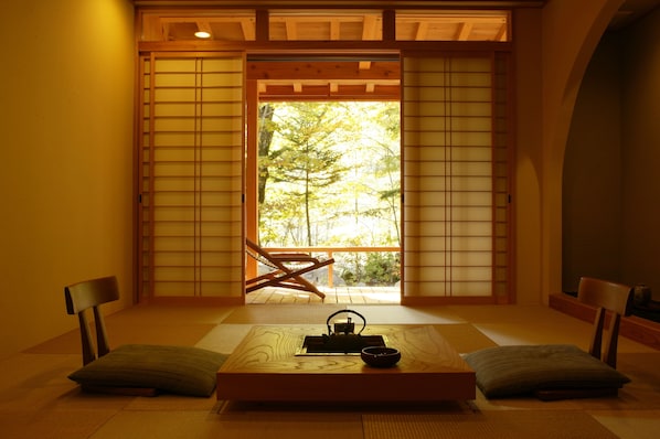 A traditional Japanese room with tatami flooring, a low table with a tea set, and sliding doors leading to a balcony with a view of trees.